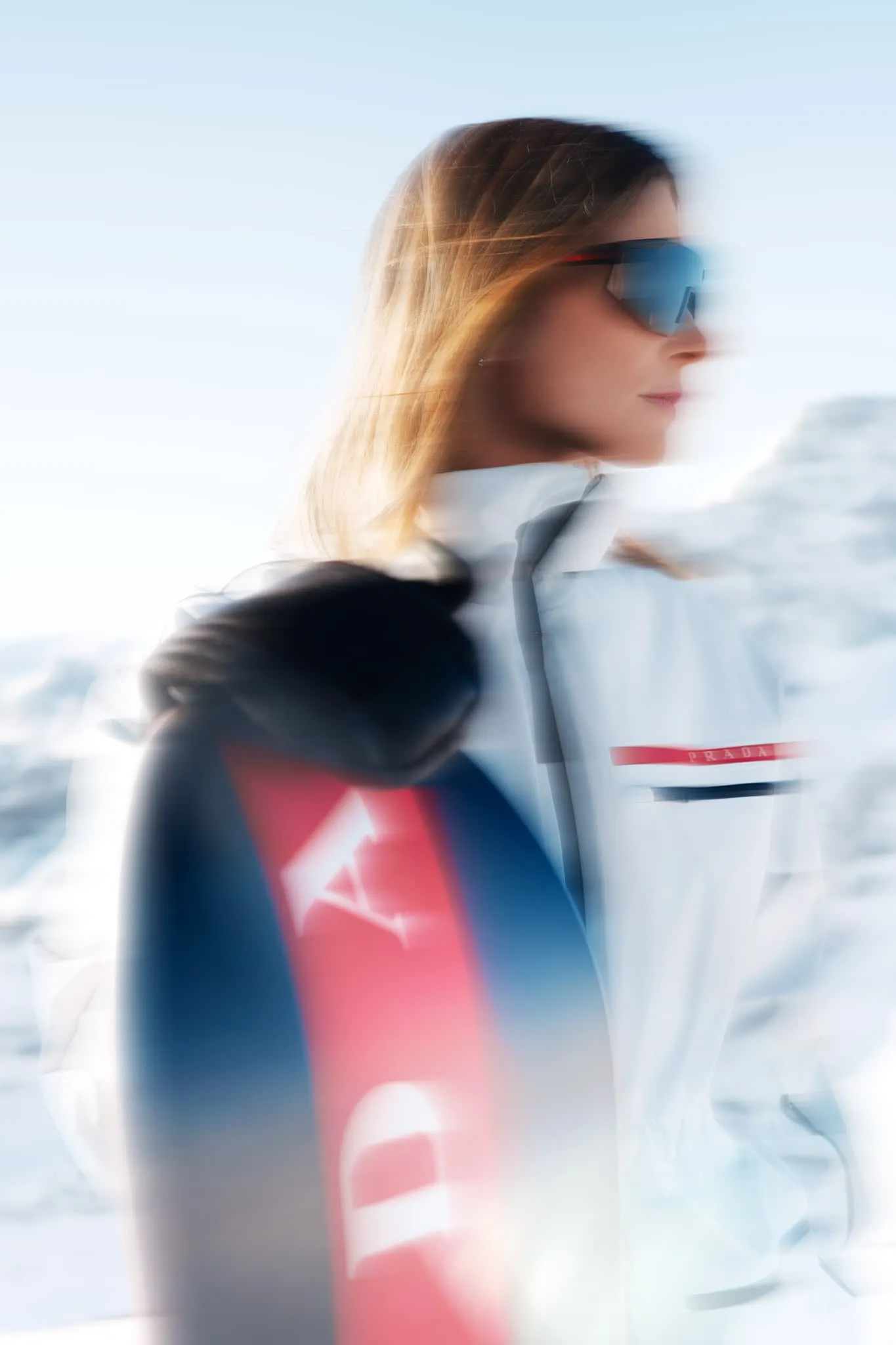A close-up, motion-blurred portrait of a woman in a white ski jacket with red and navy accents, standing against a snowy mountain backdrop. She holds a snowboard, and her long hair catches the light. The blur creates a dreamy, kinetic effect, suggesting wind and movement in cold alpine air.