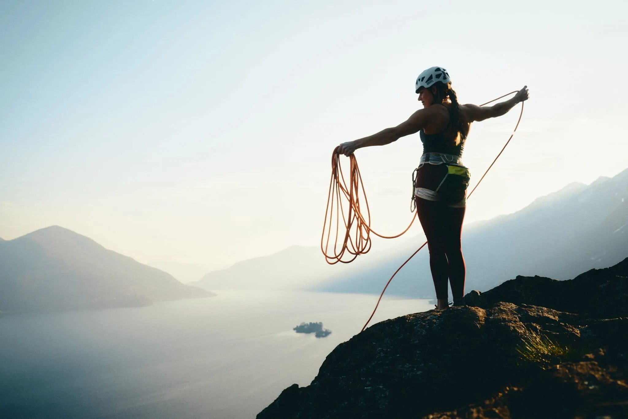 Climber Petra Klingler standing on a rocky summit at sunset, coiling a rope with Lago maggiore and mountain range in the background.