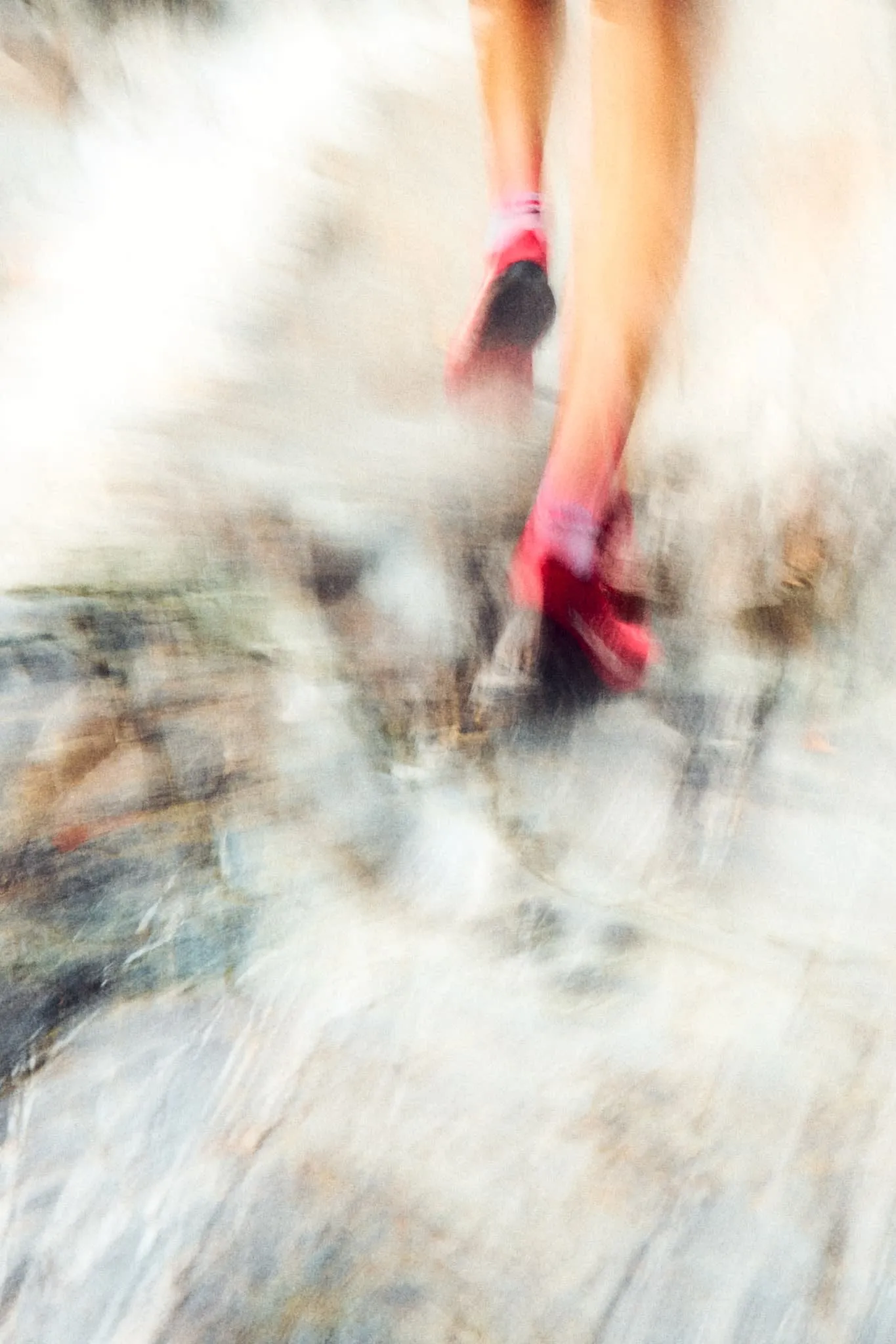 A motion-blurred image of a person running on a rocky surface, captured from behind. The runner wears bright red shoes, and the motion creates a dreamy, abstract effect with light and earthy tones.