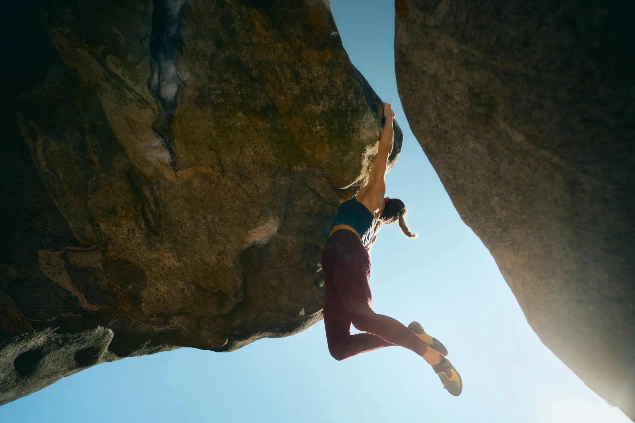 Female rock climber hangs mid-air while gripping an overhanging boulder, silhouetted against the bright sky.