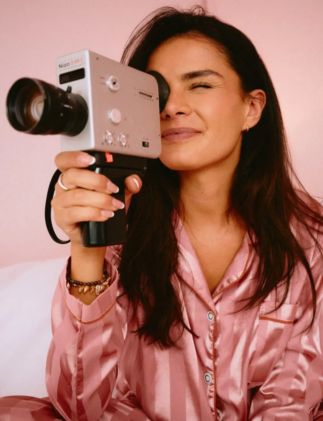  A woman in striped pink satin pajamas smiles while filming with a vintage camera, capturing the joy of creativity in a cozy, pastel-toned setting.