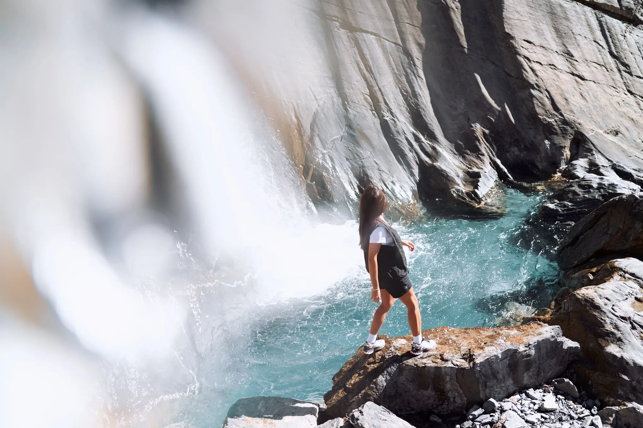 Woman standing on a rocky ledge beside a turquoise waterfall pool, gazing at the cascading water against a backdrop of smooth rock walls.