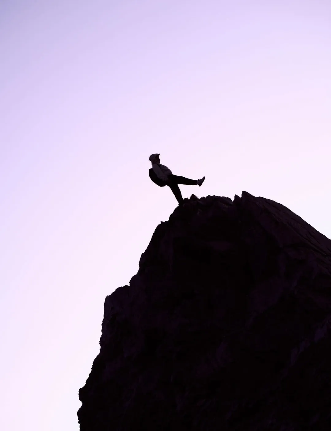 Silhouette of a hiker balancing on one leg at the summit of a rocky peak during sunset, set against a soft purple sky.