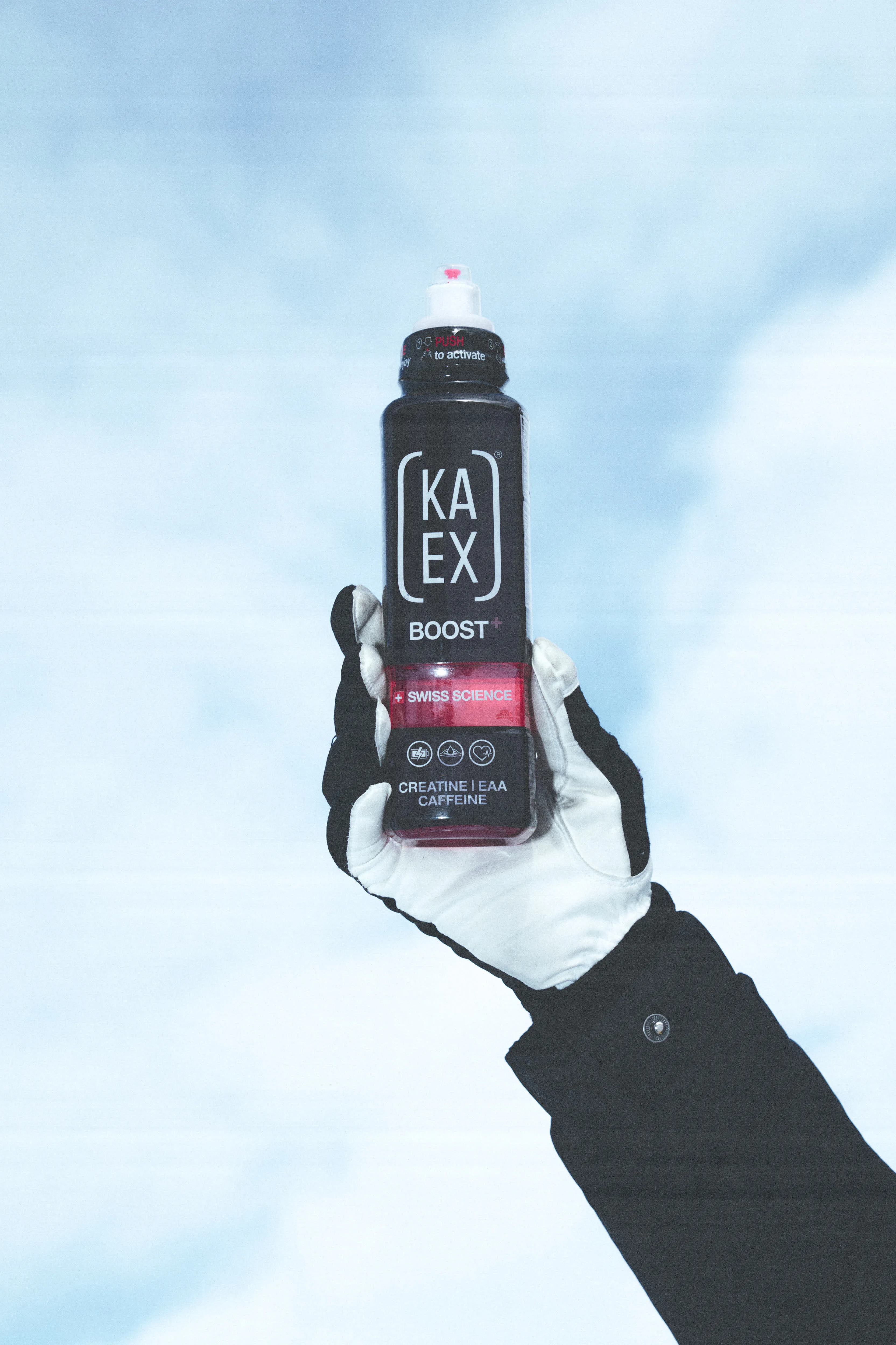 A gloved hand holds a bottle of KAEX BOOST+ supplement against a bright, clear sky. The packaging highlights "Swiss Science" and key ingredients like Creatine, EAA, and Caffeine, emphasizing athletic performance and recovery.