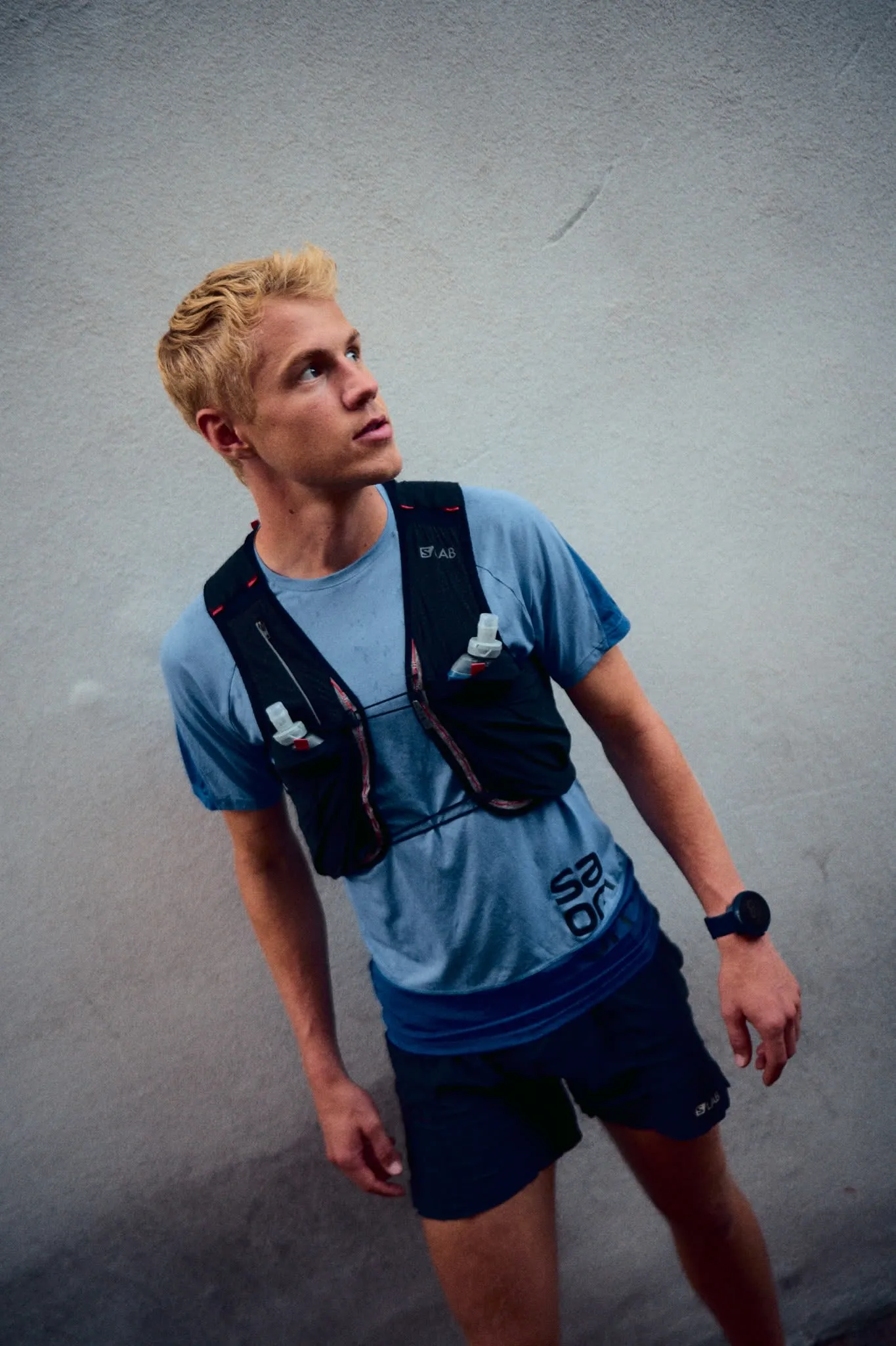 A young athletic man with blond hair stands against a light gray wall, looking slightly upward and to the left. He is wearing a light blue running shirt, dark shorts, a hydration vest, and a sports watch.