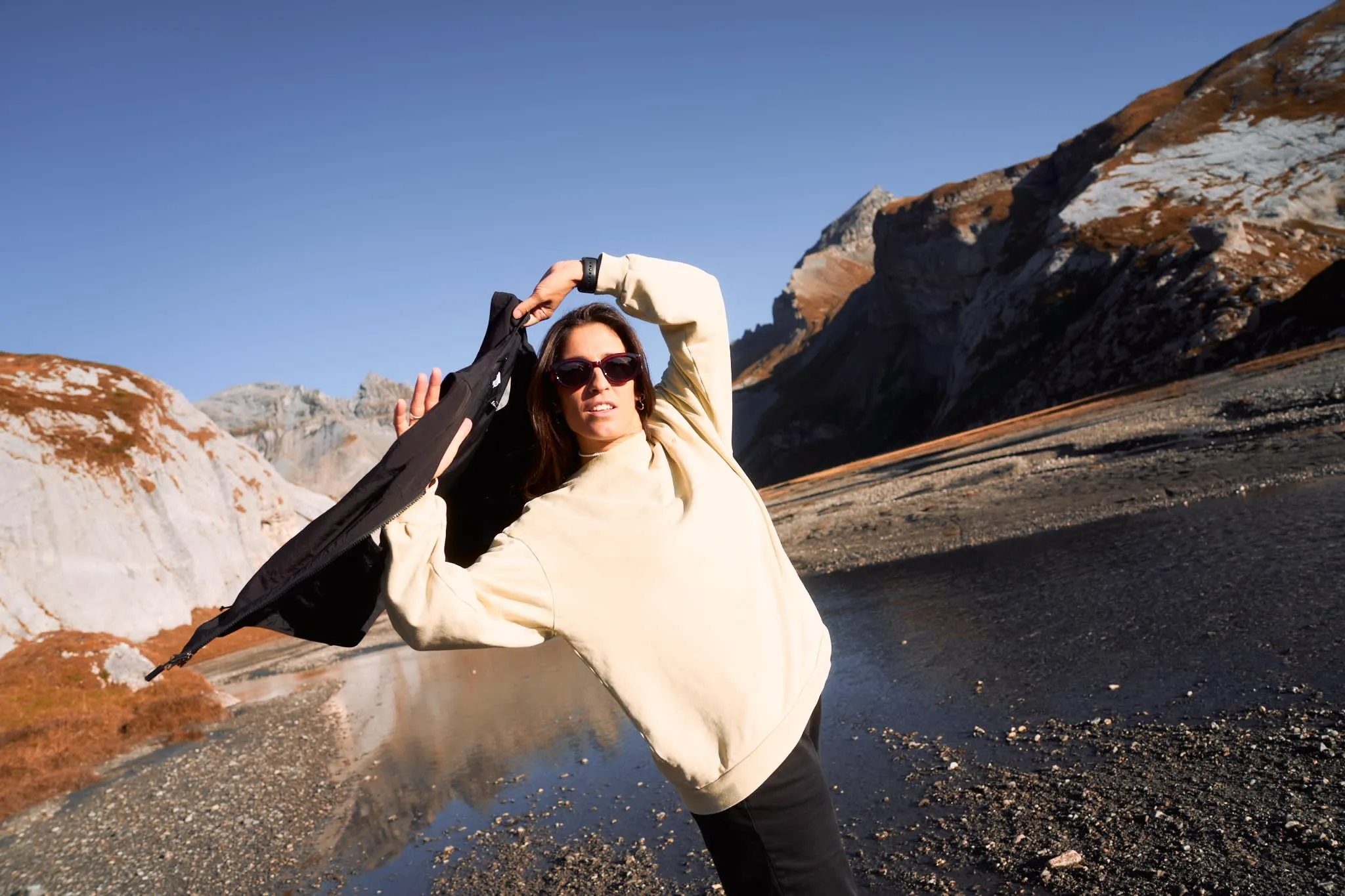 "Young woman putting on a jacket in a rugged alpine landscape, under clear blue skies, with rocky cliffs in the background."
