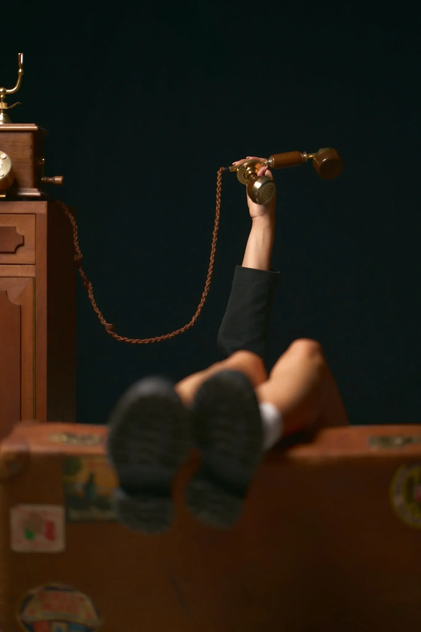 A person lying on their back with legs extended, holding up a vintage rotary telephone receiver connected by a coiled cord, with the base of the phone placed on a wooden cabinet.