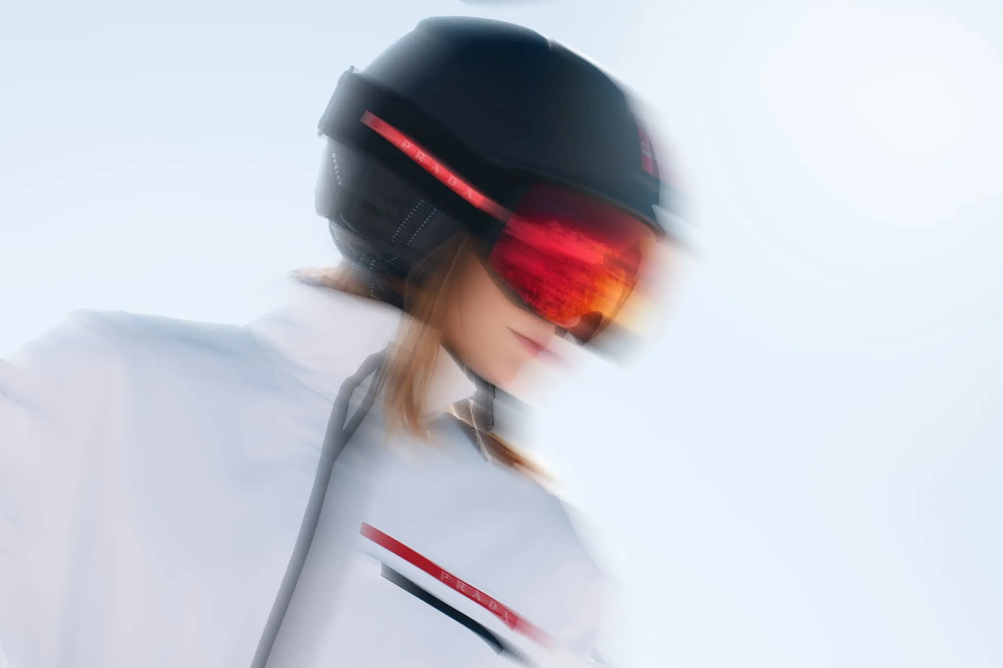A side view of a woman in a black ski helmet and red-reflective goggles, her face slightly blurred by motion. Her white jacket features red branding details, and the snowy background merges into the bright, high-key sky, enhancing the sense of speed and clarity.