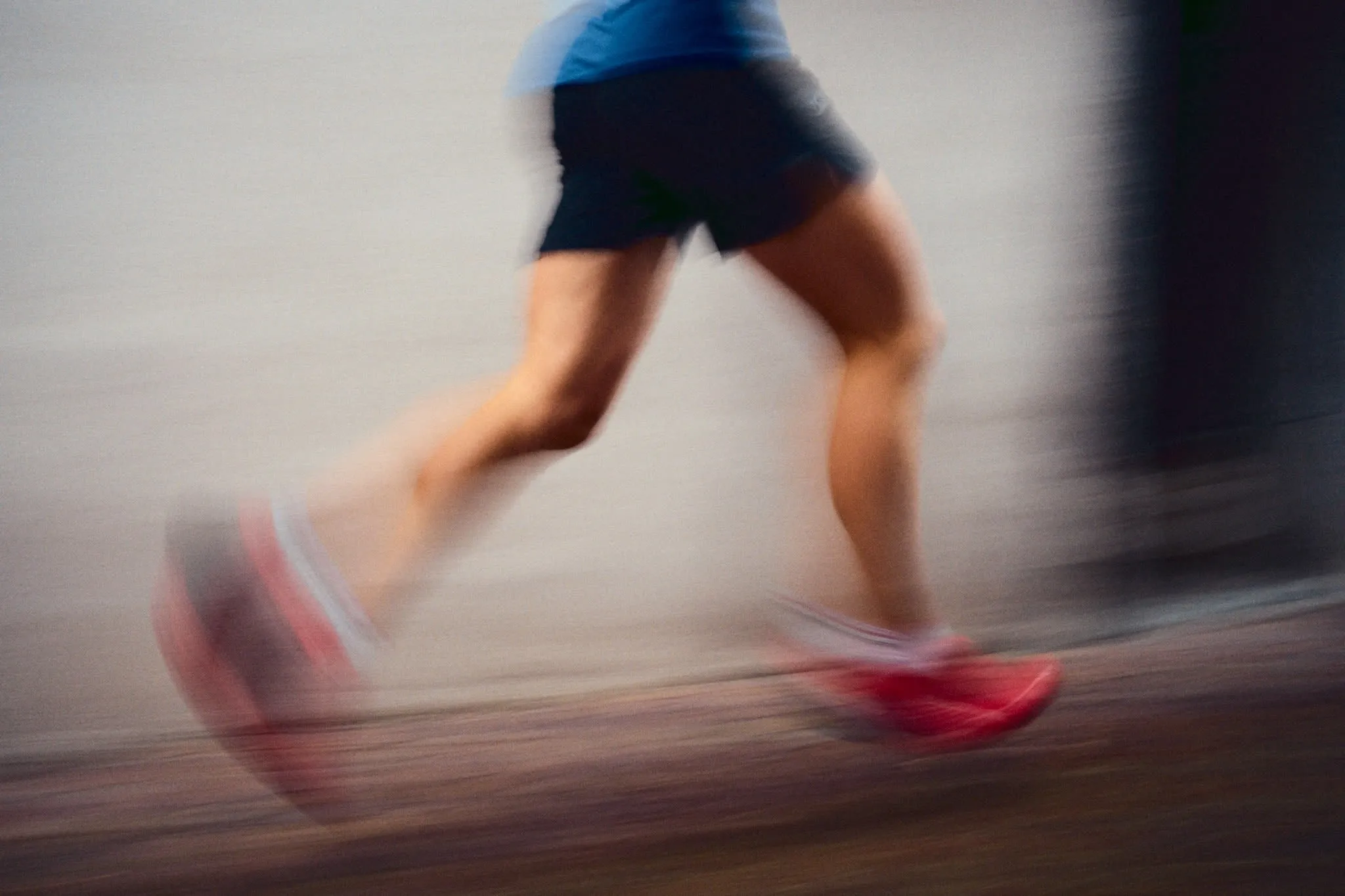 A motion-blurred image of a runner’s legs mid-stride, wearing dark shorts and bright red running shoes, moving swiftly from left to right across a smooth surface.
