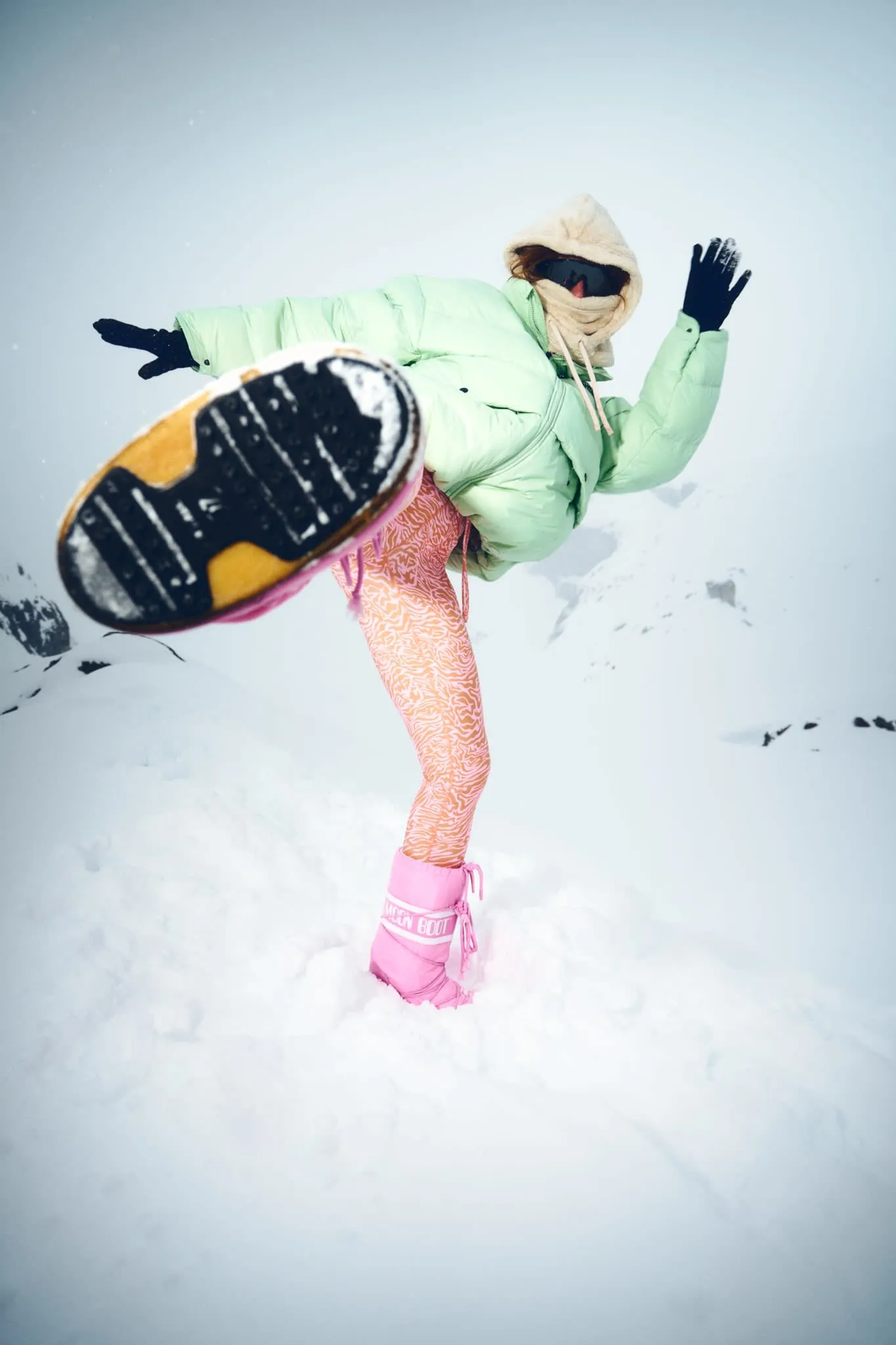 The person is caught mid-action, kicking one leg high into the air as if in a playful or dynamic pose. Their large, treaded Moon Boot is facing the camera, adding a sense of depth and motion. The snow is deep and fluffy, and snowy mountain slopes rise in the distant background under a cloudy sky. Their eccentric, colorful outfit contrasts sharply with the stark, white landscape.