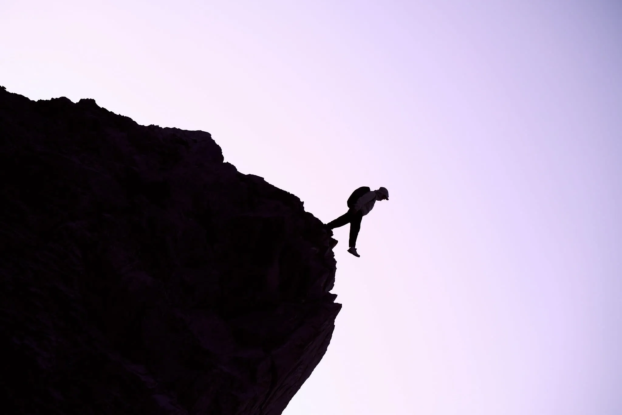 Silhouette of a hiker balancing on one leg at the summit of a rocky peak during sunset, set against a soft purple sky.