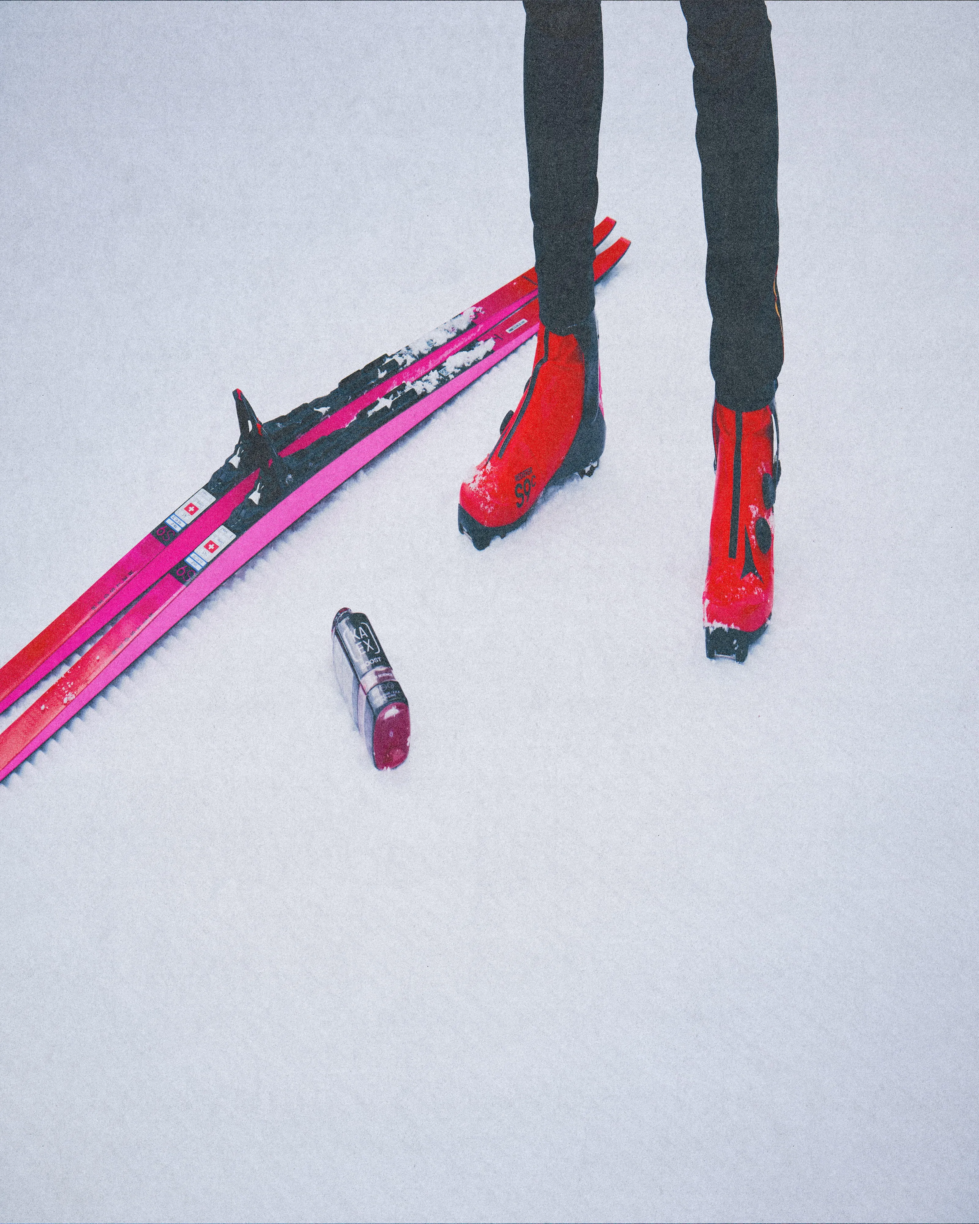 A vertical, grainy, high-angle shot featuring the lower half of a skier in red boots standing next to pink Atomic skis laid on the snow. A KAEX BOOST+ supplement bottle is positioned in the foreground, emphasizing a minimalist, high-performance aesthetic.