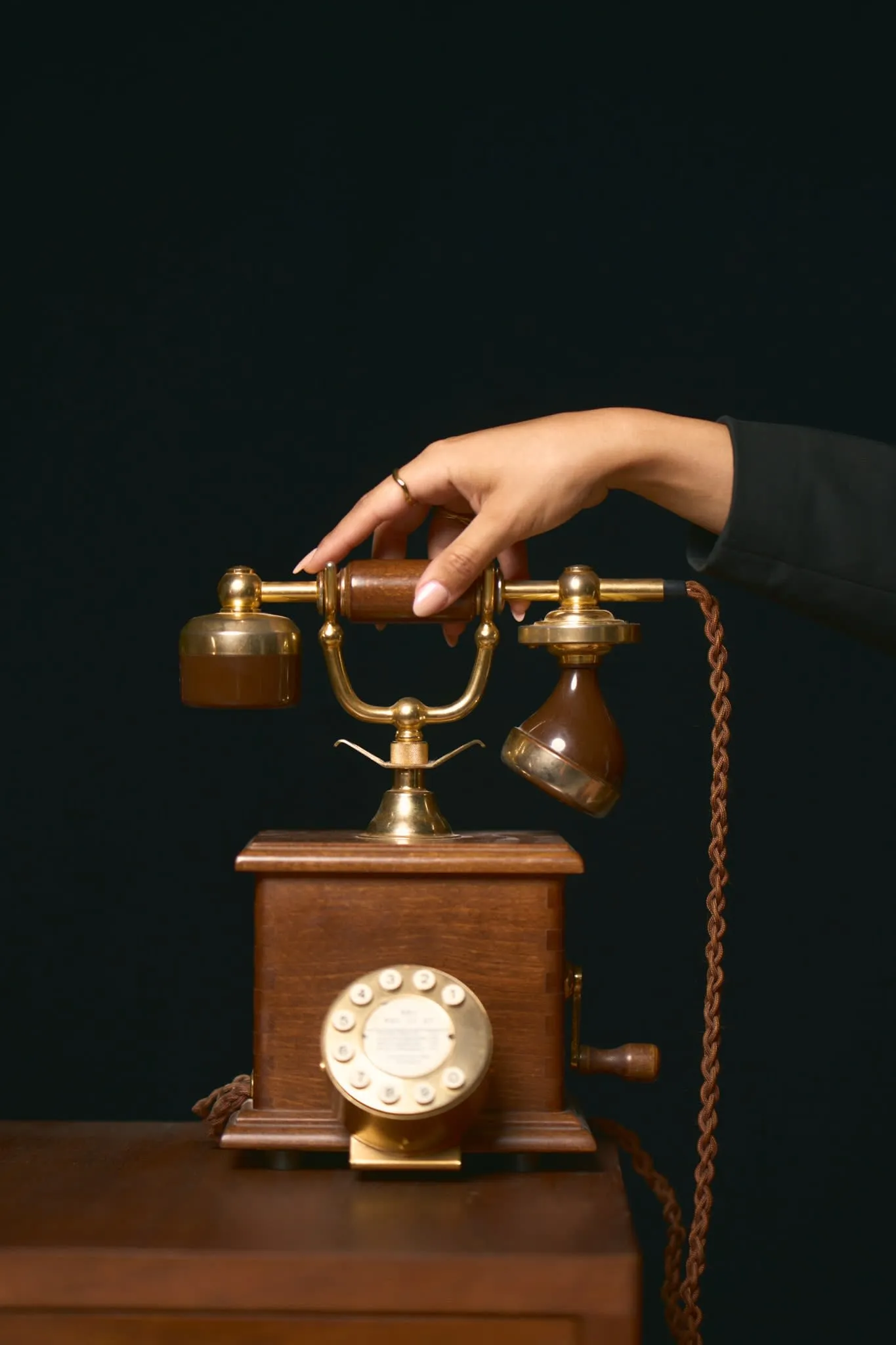 A close-up of a hand placing the receiver back on a vintage rotary telephone mounted on a wooden base, set against a dark background.
