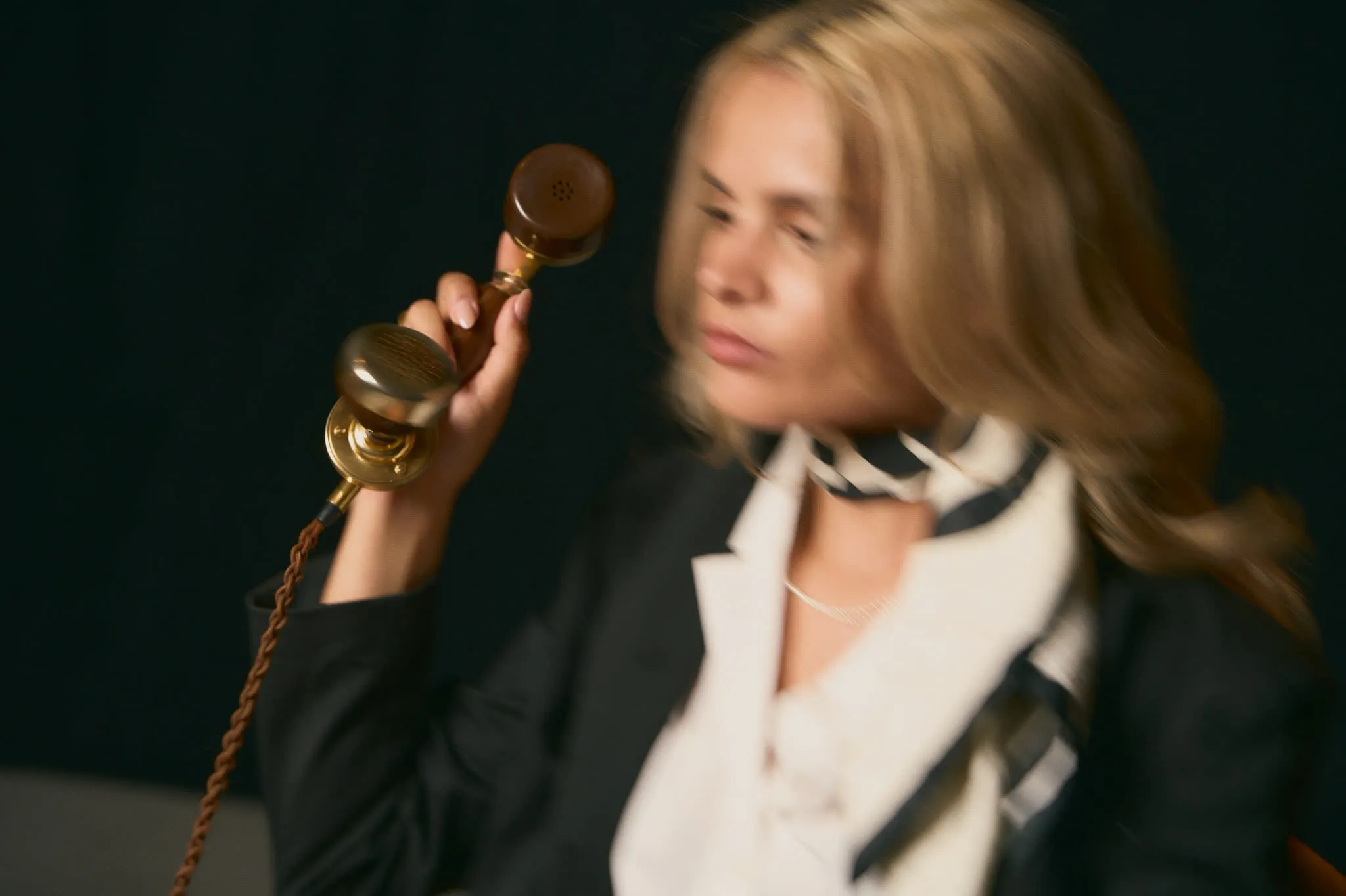 A blurred image of a blonde woman in a black blazer and striped scarf holding a vintage telephone receiver near her ear, set against a dark background.
