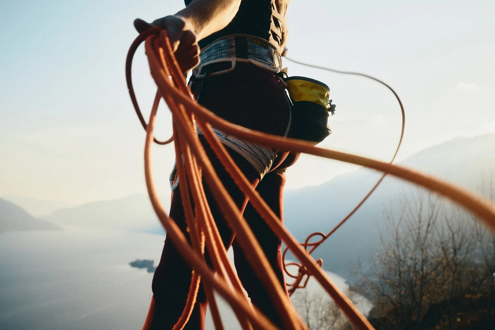Close-up of a climber’s harness and gear as they hold coiled rope, with mountains and water blurred in the background.