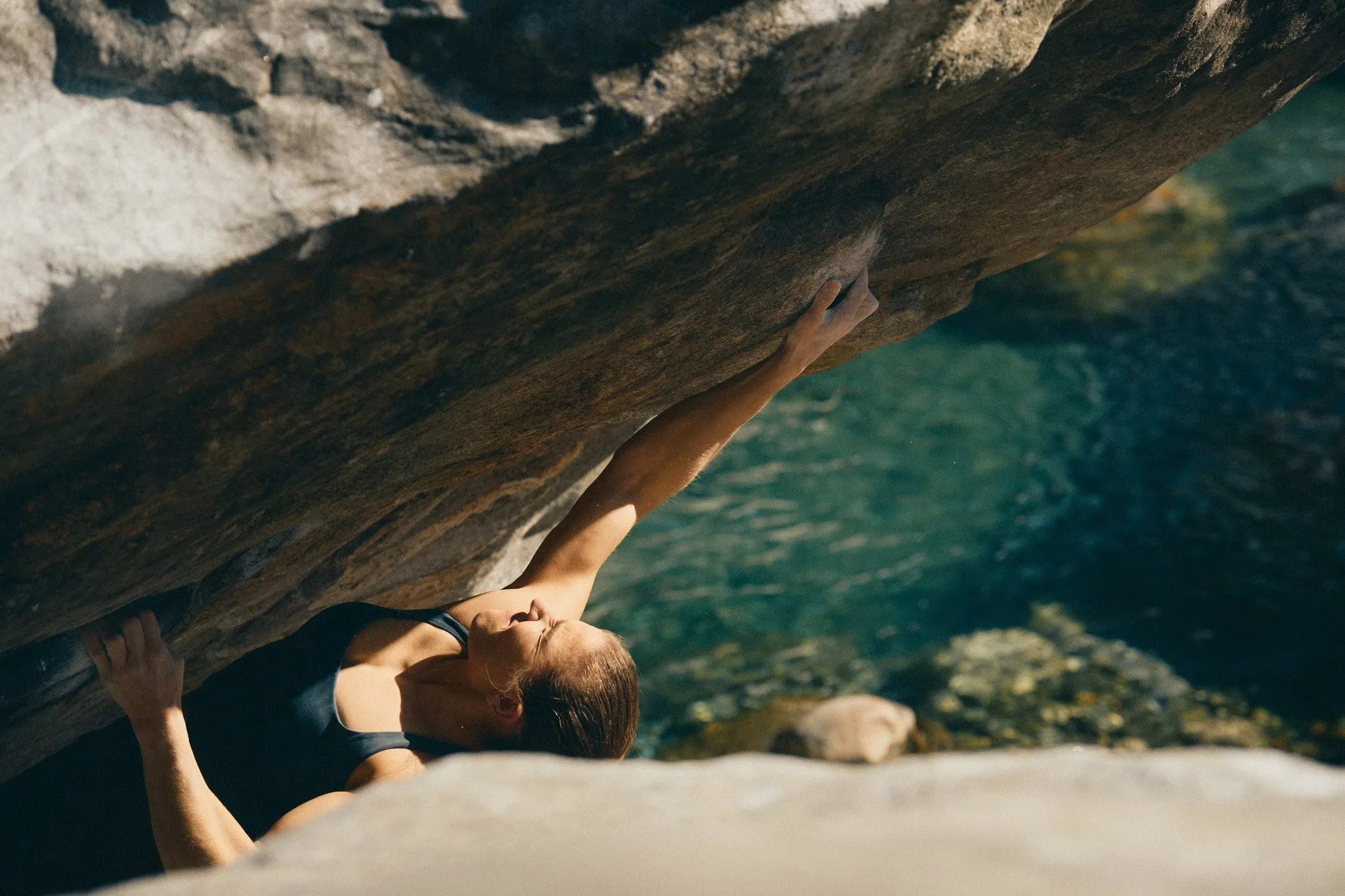 Woman scaling a slanted rock wall above clear turquoise water of val verzasca, stretching for a handhold with intense focus.