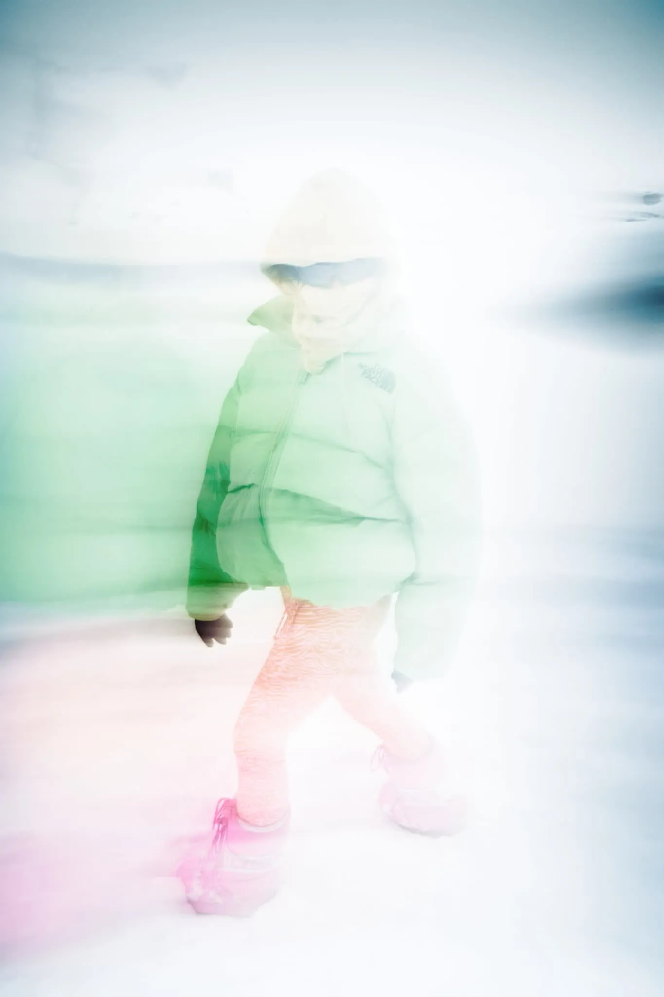 A motion-blurred photo of the same person walking through snow, wrapped in their vibrant winter attire. The image has a dreamlike, ethereal quality, with soft pastel hues blending into the snow and sky. The green jacket and pink boots are partially visible through the blur, giving a sense of movement and cold, windy conditions.
