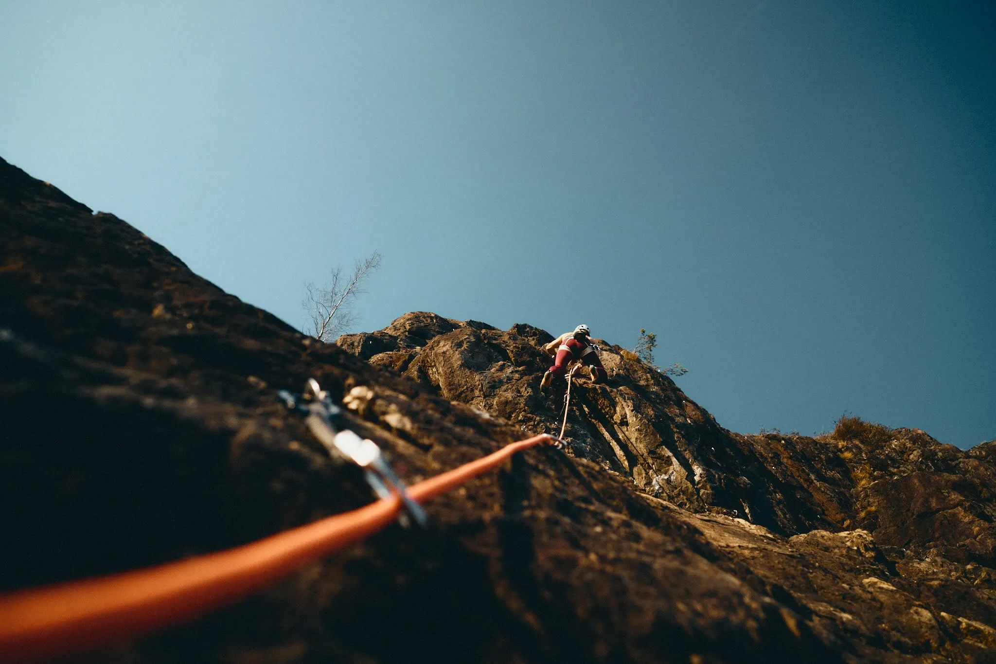 Climber ascending a steep rock face with a rope trailing below, shot from a low angle under a clear blue sky.