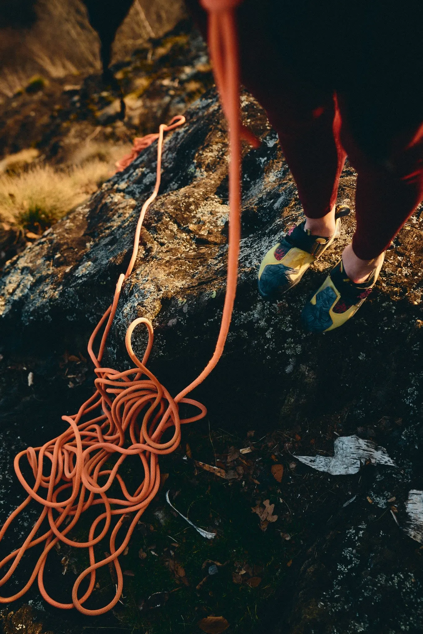View of a climber’s feet in climbing shoes next to a pile of orange rope on the rocky ground.
