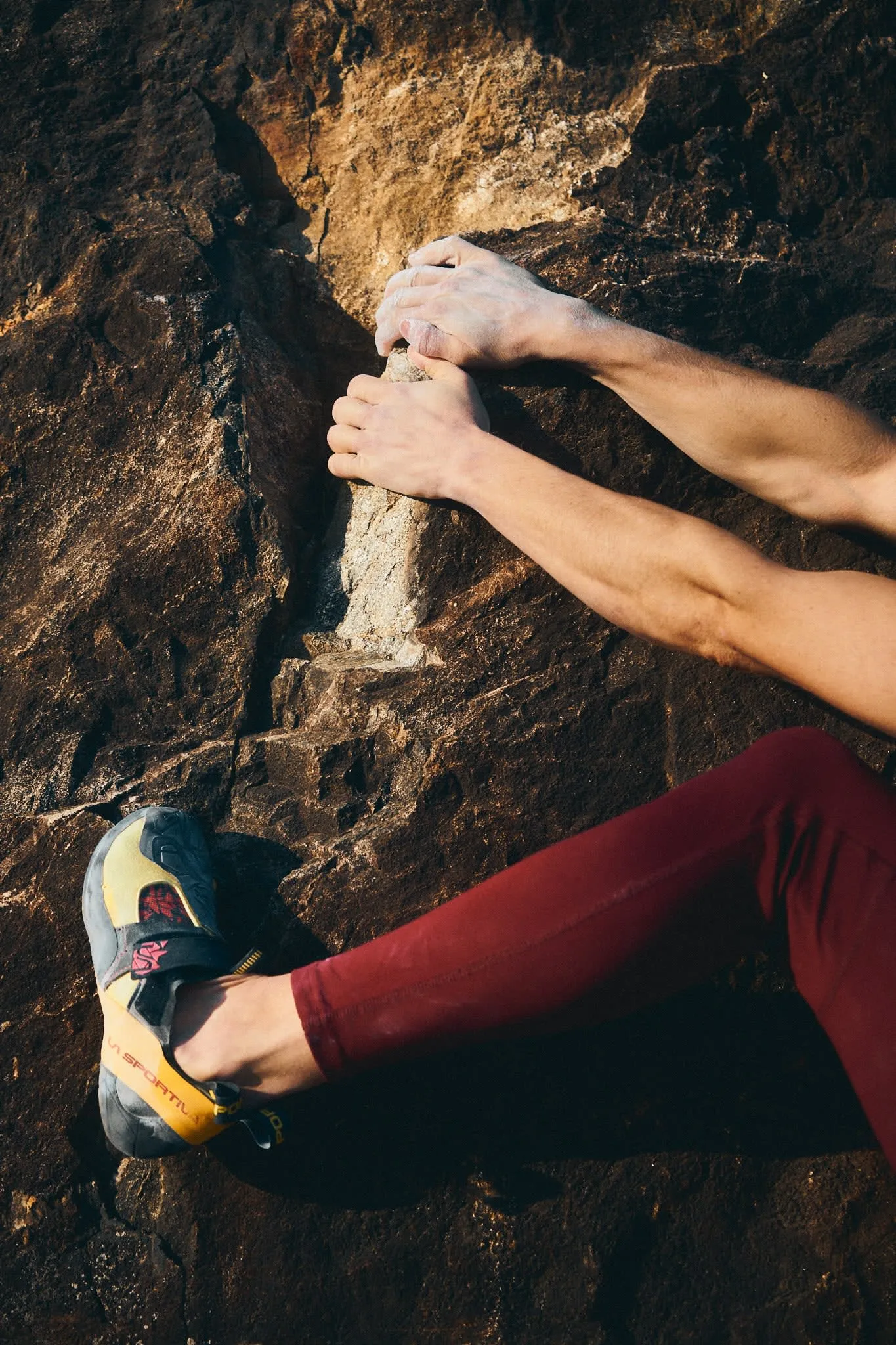 Close-up of a climber's chalk-covered hands gripping a rock crack, with one foot in a climbing shoe pressed against the wall.